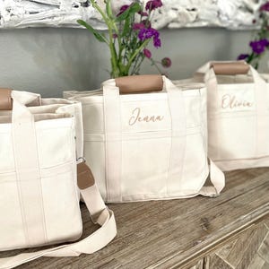 May include: Four off-white canvas tote bags with brown leather handles and straps. Each bag has a name embroidered in a light brown script: Ella, Jenna, Olivia, and another name. The bags are arranged on a wooden surface.