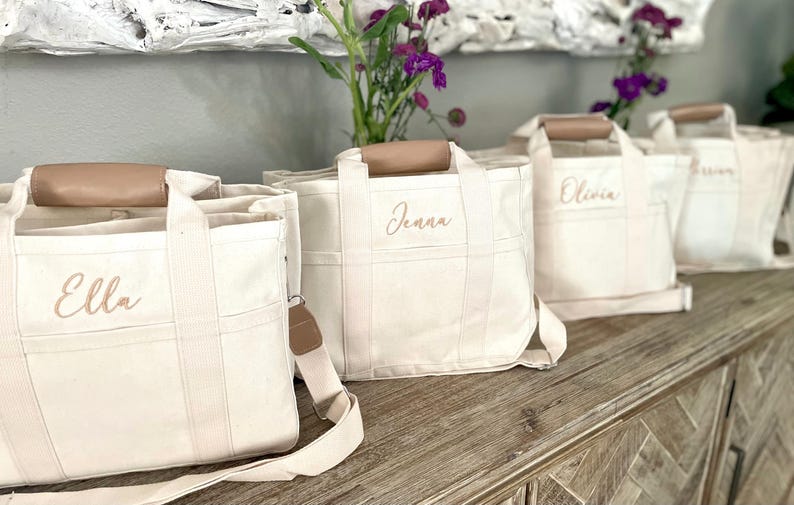 May include: Four off-white canvas tote bags with brown leather handles and straps. Each bag has a name embroidered in a light brown script: Ella, Jenna, Olivia, and another name. The bags are arranged on a wooden surface.