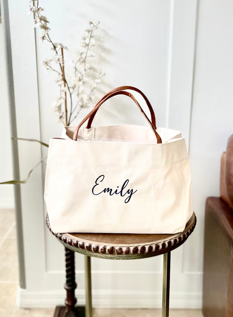 May include: A white canvas tote bag with brown leather handles. The bag has the name "Emily" embroidered on the front in black.