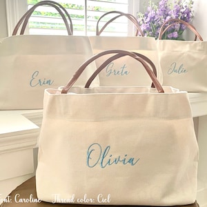 May include: Four white canvas tote bags with brown leather handles. Each bag has a different name embroidered on the front in blue thread: Erin, Greta, Julie, and Olivia. The text "Font: Caroline Thread color: Ciel" is at the bottom of the image.