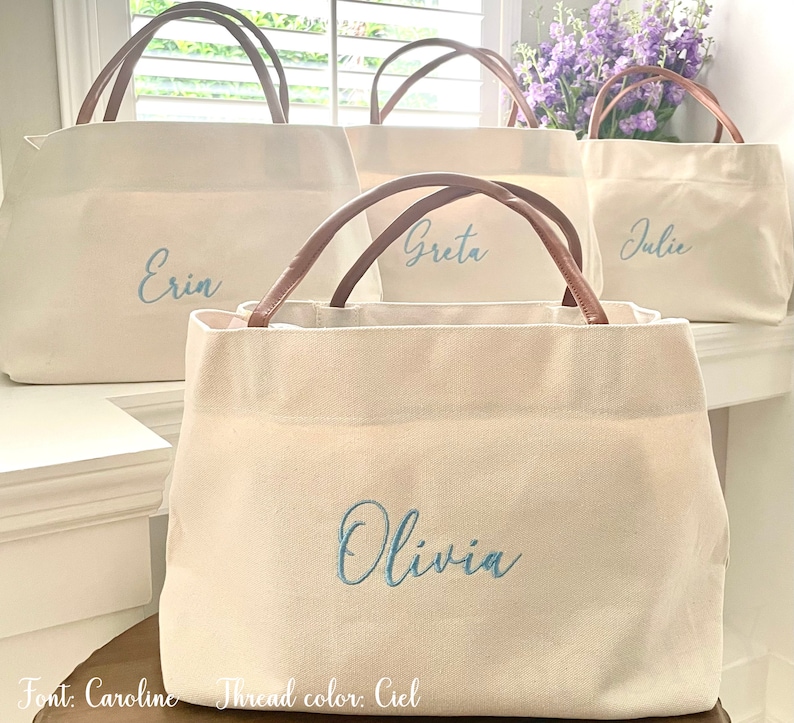 May include: Four white canvas tote bags with brown leather handles. Each bag has a different name embroidered on the front in blue thread: Erin, Greta, Julie, and Olivia. The text "Font: Caroline Thread color: Ciel" is at the bottom of the image.