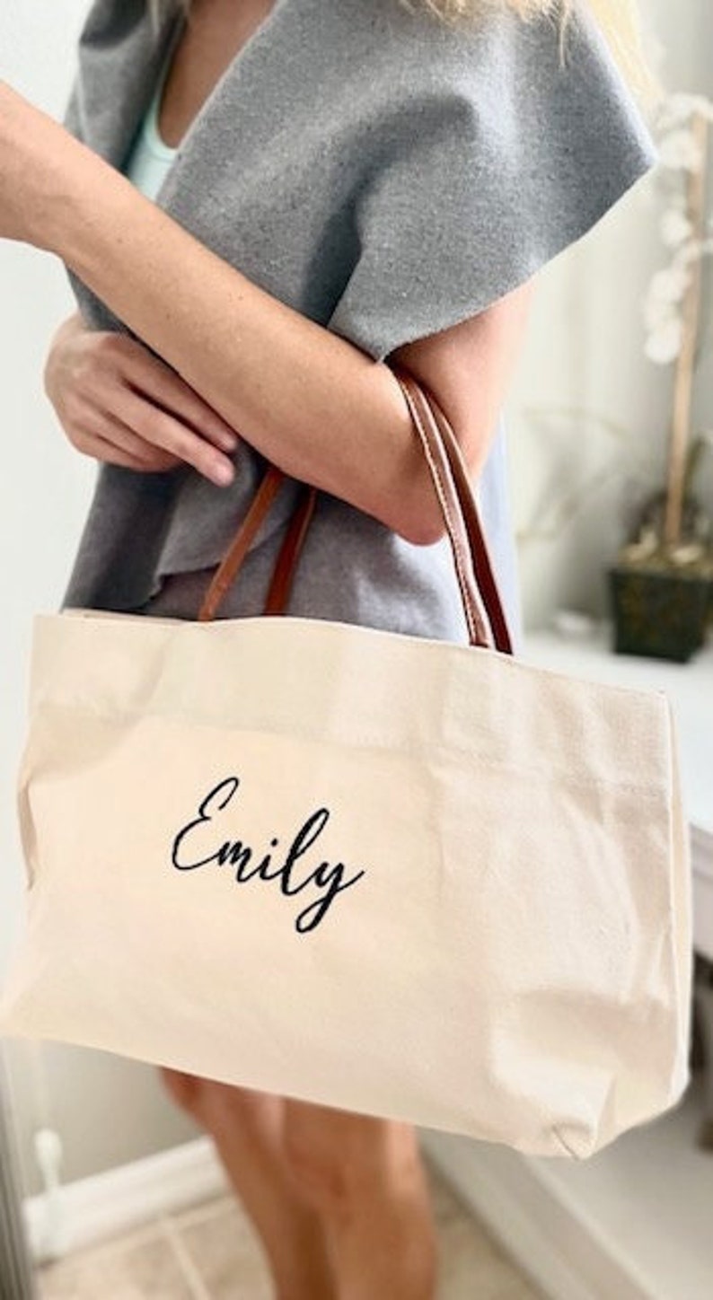 May include: Cream-colored tote bag with brown leather handles. The bag features the name "Emily" in black script. The bag is made of a sturdy canvas material, perfect for carrying everyday essentials.