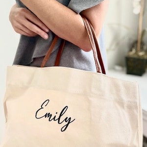 May include: A white canvas tote bag with brown leather handles. The bag has the name "Emily" embroidered on the front in black cursive.