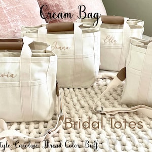 May include: Cream-colored canvas tote bags with brown leather handles and personalized names embroidered in buff thread. The bags have exterior pockets and adjustable shoulder straps. Text on the image reads "Cream Bag" and "Bridal Totes."