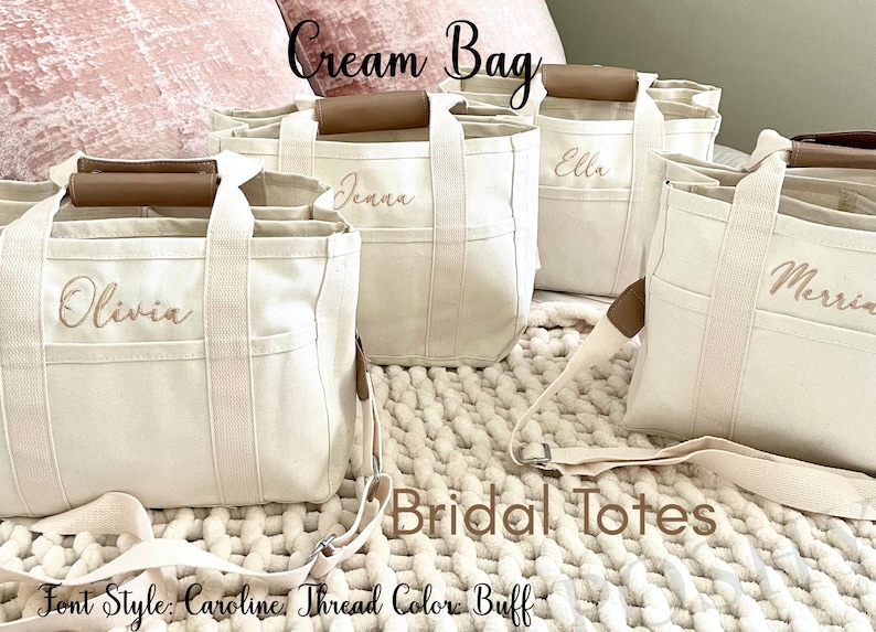 May include: Cream-colored canvas tote bags with brown leather handles and personalized names embroidered in buff thread. The bags have exterior pockets and adjustable shoulder straps. Text on the image reads "Cream Bag" and "Bridal Totes."