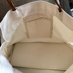 May include: A white canvas tote bag with brown leather handles and a zippered pocket on the inside. The bag is empty and the lining is visible.
