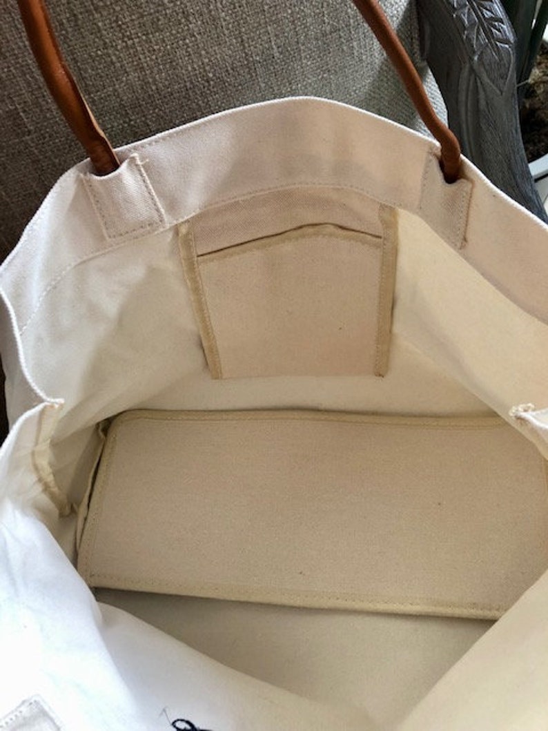 May include: A white canvas tote bag with brown leather handles and a zippered pocket on the inside. The bag is empty and the lining is visible.