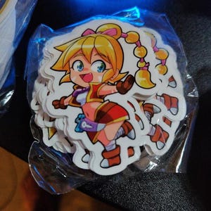 May include: A stack of stickers featuring a cartoon girl with blonde hair, blue eyes, and a cheerful expression. The girl is wearing a red and brown outfit. The stickers are in a clear plastic bag.