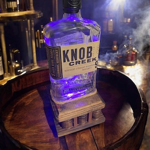May include: A clear glass bottle of Knob Creek Kentucky Straight Bourbon Whiskey, aged nine years, sits on a wooden stand. The bottle is illuminated with blue light, and the label is visible. The stand is made of wood and has a decorative design.