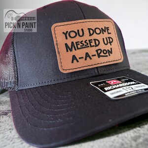 May include: Black trucker hat with a brown leather patch that says "You Done Messed Up A-A-Ron".