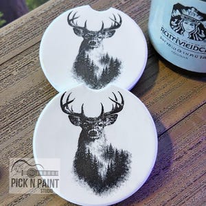 May include: Two white car coasters with a black and white deer and forest design. The coasters have a small notch for easy removal. The text "Pick N Paint Studio" is visible on one coaster.