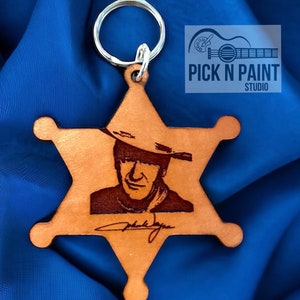 May include: A brown leather sheriff's star keychain with a laser engraved image of John Wayne wearing a cowboy hat and a signature. The keychain is attached to a silver key ring.