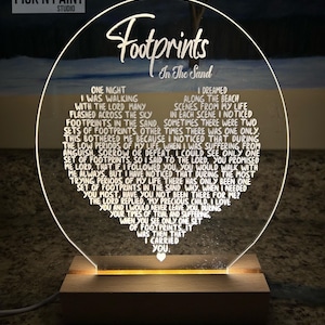 May include: A white acrylic plaque with a heart-shaped cutout, illuminated from behind. The plaque features the poem "Footprints in the Sand" with a handwritten font. The poem tells a story about a person walking with God and seeing only one set of footprints during difficult times. The plaque is mounted on a wooden base.