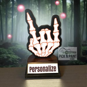 May include: A black and white skeleton hand sign with orange accents, mounted on a wooden base. The base has a light-up panel with the word "Personalize" in white. The background is a blurred forest scene.
