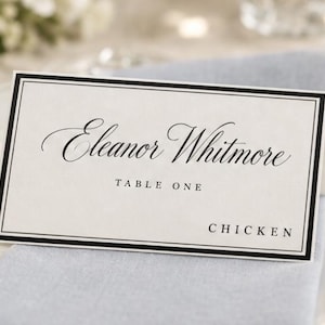 May include: A rectangular white folded place card with a double black border. The card features the printed name Eleanor Whitmore in a black script font, with the text TABLE ONE and CHICKEN printed in a simple black sans-serif typeface below.