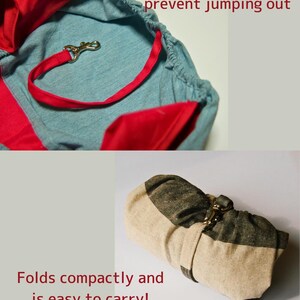 Op de afbeelding: Een lichtblauwe en rode stoffen baby draagzak met een bevestigingsband en een metalen sluiting. De draagzak is opgevouwen en vastgemaakt met de band. De tekst "A tether strap to prevent jumping out" en "Folds compactly and is easy to carry!" is zichtbaar in de afbeelding.