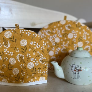 May include: Two yellow tea cozy covers with a white floral pattern. The covers are on a white surface with a green teapot with white polka dots and a sheep illustration.