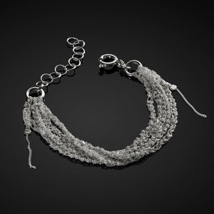 May include: A silver bracelet with multiple strands of delicate, shimmering chains. The bracelet has a chain extension and a secure clasp. The design is elegant and suitable for various occasions. The bracelet is displayed against a dark background.