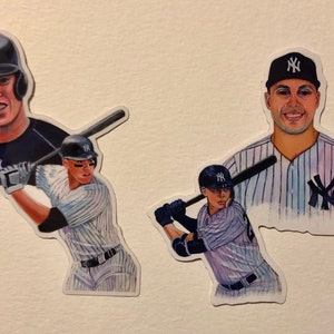 May include: Three watercolor illustrations of baseball players wearing New York Yankees uniforms. The first illustration shows a player in a blue jersey with a white 'NY' logo, the second shows a player in a pinstripe jersey with a white 'NY' logo, and the third shows a player in a pinstripe jersey with a white 'NY' logo.