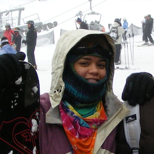 May include: A woman wearing a white hooded jacket with a colorful scarf and a black face mask. She is holding a black snowboard.