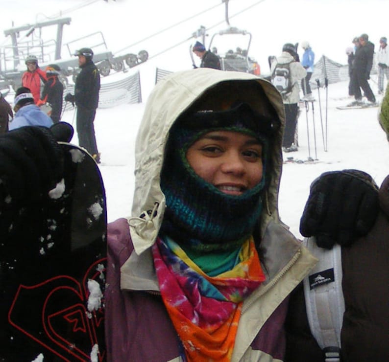 May include: A woman wearing a white hooded jacket with a colorful scarf and a black face mask. She is holding a black snowboard.
