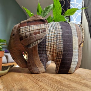 May include: A stuffed elephant figurine made from various brown tweed fabrics. The elephant has large ears, a trunk, and a small tail. The fabrics include plaid, herringbone, and solid patterns. The elephant is sitting on a wooden surface.
