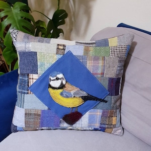 May include: A bluebird embroidered on a patchwork cushion with a blue background. The cushion is made of various fabrics in shades of blue, brown, and beige.