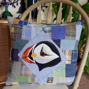 May include: Square patchwork pillow with a colorful design. The pillow features a central square with an embroidered puffin bird design. The patchwork consists of various fabrics in shades of blue, green, gray, and brown.