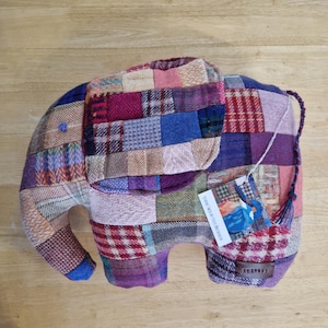 May include: A patchwork elephant-shaped pillow in shades of purple, blue, pink, and brown. The pillow has a tag attached with text and a tassel tail. The elephant's trunk and feet are a solid blue color.