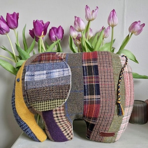 May include: A patchwork elephant toy made from different fabrics, including plaid and tweed. The elephant is standing on a white surface with a bouquet of purple tulips in the background.