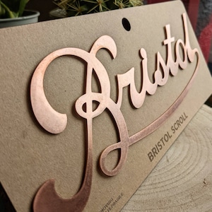May include: A copper-colored metal sign with the word "Bristol" in a stylized script font. The sign is on a brown background and has the text "Bristol Scroll" printed below the word "Bristol".