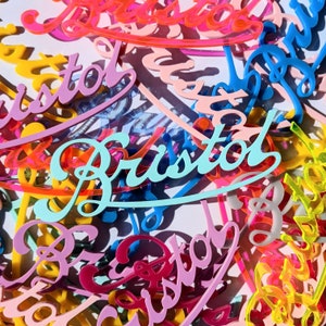 May include: A colourful assortment of acrylic signs with the word "Bristol" in blue and white lettering. The signs are in various colours, including pink, yellow, orange, and green.