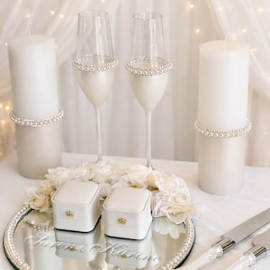 May include: Elegant wedding decor featuring two champagne flutes adorned with pearl accents, two white ring boxes, and two pillar candles. A mirrored tray with floral accents and the names "Karen & Karine" is also present.