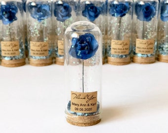 May include: A collection of glass vials, each containing a blue rose and glitter. The vials have cork stoppers and small labels with the text "Thank You" and a date. The roses are a deep blue color.
