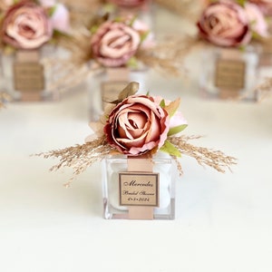 Może przedstawiać: Przezroczyste pudełka na prezenty z akrylu ozdobione pojedynczą różową różą i suszoną trawą. Pudełka zawierają białe cukierki i mają spersonalizowaną etykietę z napisem "Mercedes Bridal Shower 6-8-2023".