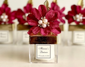 May include: Small clear glass jars with gold lids and a burgundy flower with pearl accents on top. Each jar has a white label with the text "Iliana's Quince" and "November 8, 2021".