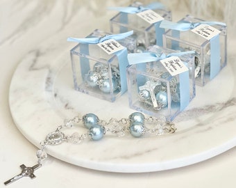 Puede incluir: Cuatro cajas de plástico transparente con cinta azul y una etiqueta blanca con el texto "Liam's Baptism July 2, 2023". Cada caja contiene un pequeño rosario azul. Un rosario plateado con cuentas azules está sobre una superficie de mármol blanco.
