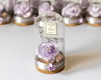 May include: A small glass dome with a cork base contains a purple satin flower, silver beads, and a small tag with the text "Leydricha & Kushal 6.20.20".