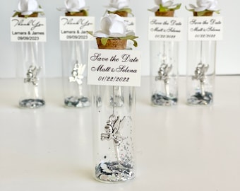 May include: A set of six small glass bottles with cork tops, each containing silver glitter and a silver fairy charm. The bottles are decorated with white flowers and small cards with the text "Save the Date" and the names "Matt & Selena" and the date "01/22/2022".