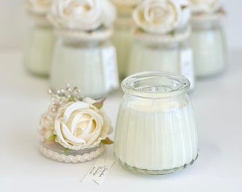 May include: Set of small glass jars with white candles and white flower accents. The jars have a ribbed texture and are decorated with a small white flower on top. The jars are arranged in a row on a white surface.