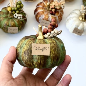 May include: A collection of small, decorative pumpkins with various paint finishes and embellishments. The pumpkins are decorated with fall-themed accents, such as berries, leaves, and twine. One pumpkin is held in a hand and has a small tag that reads "Thankful".