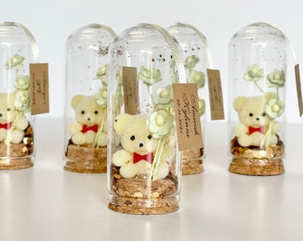 May include: Five clear glass jars with cork lids contain a small plush teddy bear, a sprig of green flowers, and gold glitter. A brown tag with the text "Sophia Kylie 12-11-2021" is attached to one of the jars.