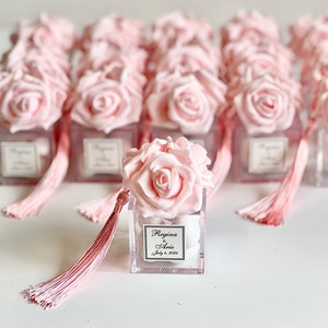 以下が含まれることがあります： ピンクのバラの花の飾り付けが施された透明な正方形の箱に、ピンクのタッセルが付いています。箱には、「Regina & Arie 2023年7月8日」という白いラベルが付いています。