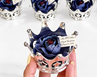 May include: A silver crown-shaped container holding a dark blue fabric flower with a small white tag printed with the text Mis XV Años, Anjali Contreras, August 17, 2024. The crown features a metallic finish and intricate cut-out details.