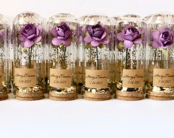 May include: A row of small glass jars with cork tops filled with gold glitter and a single purple paper flower. Each jar has a brown paper label with the text "Henry & Tamara 2.26.2021".