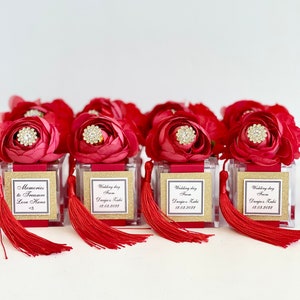 Puede incluir: Cajas de regalo de boda rojas y doradas con una flor roja y un strass en la parte superior. Las cajas tienen un frente transparente y un fondo de purpurina dorada. Las cajas tienen un borla roja que cuelga de la parte inferior. Las cajas tienen una etiqueta con el texto "Wedding day from Dunja & Kaki 12.03.2022".