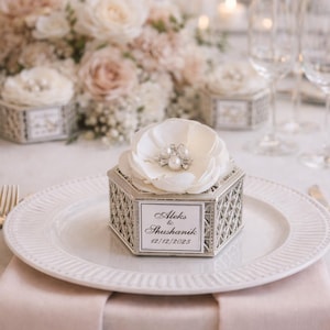 May include: A silver geometric box, topped with a white flower and pearl embellishments, rests on a white plate. The box has a label with the names "Aleks & Shushanik" and the date "12/12/2025". The setting includes a pink tablecloth and floral arrangements.