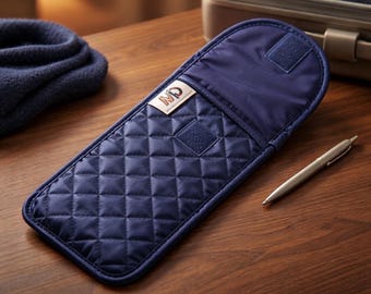 May include: A navy blue quilted fabric case with a rounded top flap and a hook-and-loop closure. The case features a small rectangular label with the letters ND printed in orange and black. A silver metal pen is positioned next to the case.