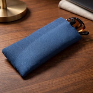 Puede incluir: Una funda rectangular de tela azul con un tejido texturizado, que contiene un par de gafas con montura marrón parcialmente insertadas. La funda tiene un diseño plano y sencillo, confeccionada con un material textil liso con costuras visibles.