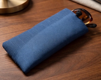 May include: A rectangular blue fabric pouch with a textured weave, featuring a pair of brown-rimmed glasses partially tucked inside. The pouch has a simple, flat design and is constructed from a solid-colored textile material with visible stitching.
